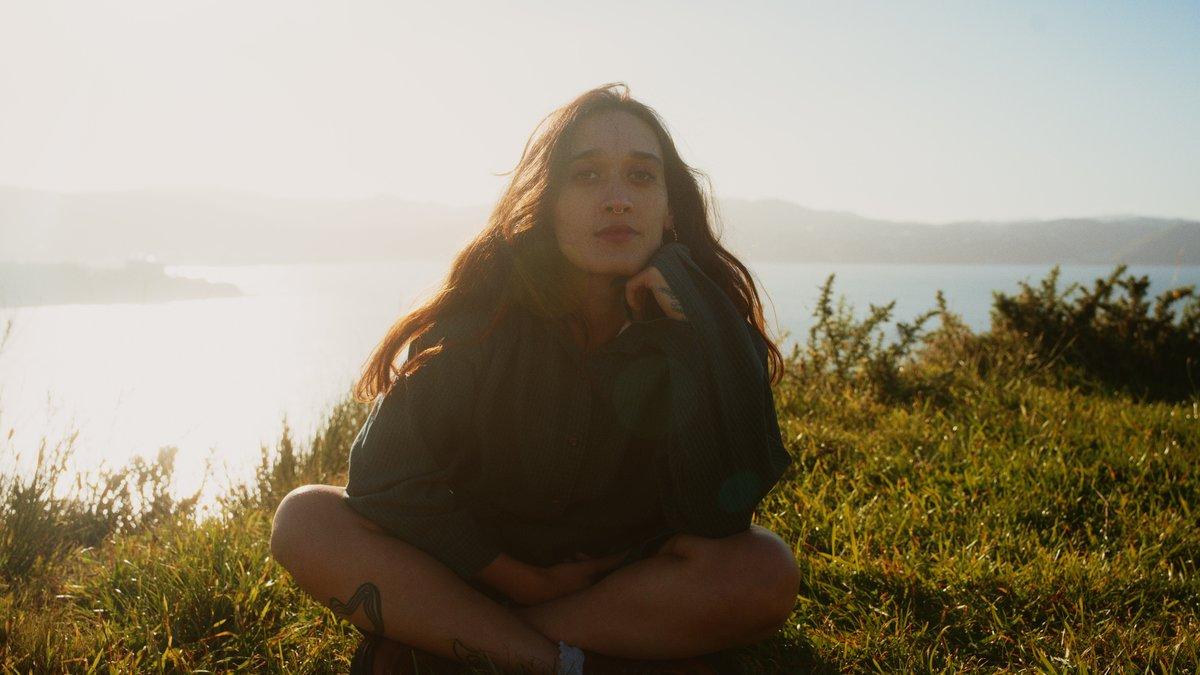 Lily sits thoughtfully on a hilltop, with a dramatic and dreamy landscape of oceans and hills in the background