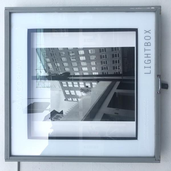 Image of lightbox at Thistle hall in daylight with Black and white image of young woman reaching out to the photographer in a gesture of friendship. Sun burst on window of building behind subject. 