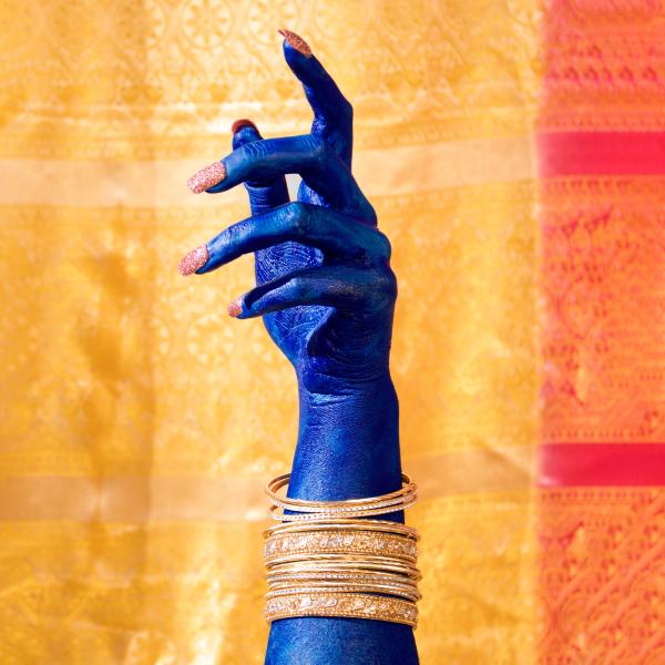 Woman's hand and lower arm painted bright blue with long pink glittered nails. Multiple gold bangles. Gesture of hand is loosely pointing upwards and is set against a background of gold and red fabric.