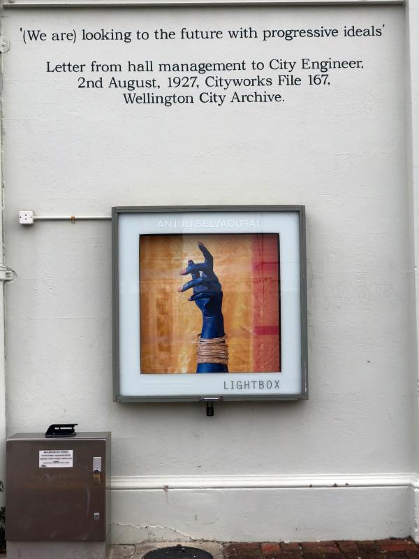 Install shot of artwork in the lightbox at thistle hall - Woman's hand and lower arm painted bright blue with long pink glittered nails. Multiple gold bangles. Gesture of hand is loosely pointing upwards and is set against a background of gold and red fabric.