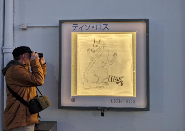 Tiso Ross artwork 'Bikurishita' installed in Thistle Hall Lightbox 3 August 2025. A large drawing of a dragon like creature. It is perched on a pillow, jumping up in suprise. The drawing is lit up in the evening gloom.