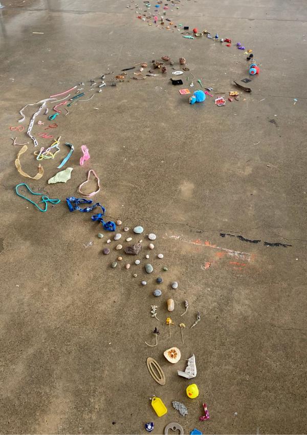 Sculptural installation by Morgan Dean. A line of small loss and found objects snakes across a concrete floor.
