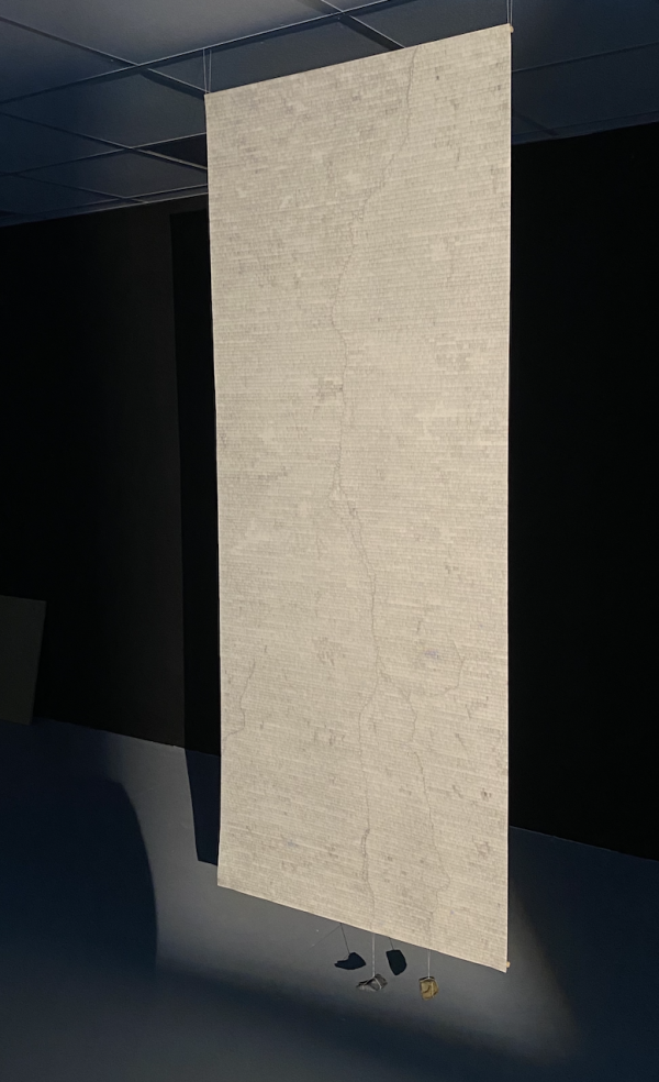 A monolithic-looking rectangle hangs from the ceiling in a dimly-lit room, with small objects hanging off the bottom of it